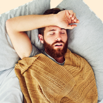 Male adult sleeping with two pillows behind his head and a brown blanket to cover him.