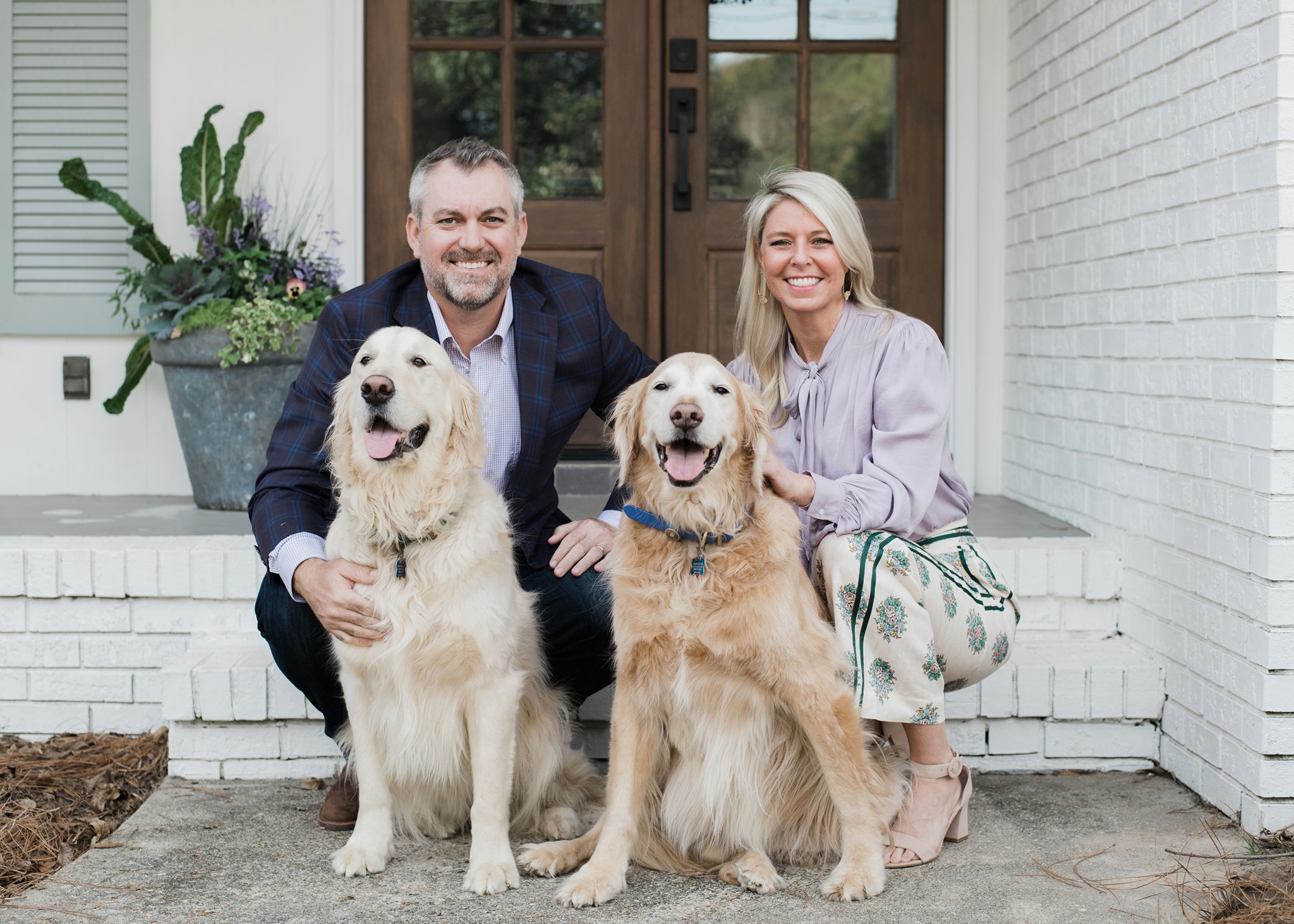 A picture of Dr. Gordon & kelly smiling while sitting in the front of their home and petting their two gold retriever dogs.
